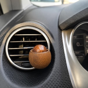 Puede incluir: Un clip ambientador esférico para coche sujeto a una rejilla de ventilación circular del salpicadero. El dispositivo presenta una carcasa exterior de color marrón mate y textura rugosa con una cúpula ámbar lisa y brillante en el centro.