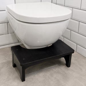 May include: A black wooden toilet stool with a rectangular top and four legs, positioned directly in front of a white ceramic toilet. The stool features a curved cutout on the side facing the toilet base and is secured with visible black screws.