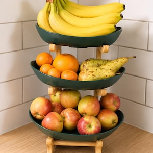 May include: A three-tiered fruit stand featuring dark green, oval-shaped bowls supported by a light-colored wooden frame. The top bowl holds a bunch of yellow bananas, the middle bowl contains oranges and pears, and the bottom bowl holds red apples.