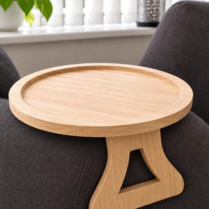 May include: A light-coloured wooden armrest tray featuring a circular top surface with a raised edge and a sturdy, downward-extending support leg with a triangular cutout, designed to rest securely over the arm of a piece of furniture.