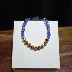 May include: A beaded bracelet featuring alternating blue and bronze faceted crystal beads. The centre of the bracelet displays round metallic beads with the black embossed letters spelling out the word RAVENCLAW in a circular arrangement.