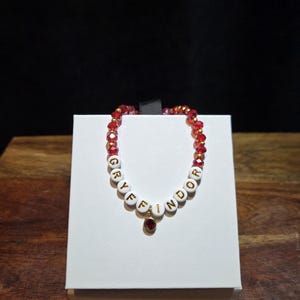 May include: A beaded bracelet featuring red faceted crystals and white round beads with gold lettering that spells out GRYFFINDOR. The bracelet includes a small circular charm with a red stone hanging from the center of the lettered beads.