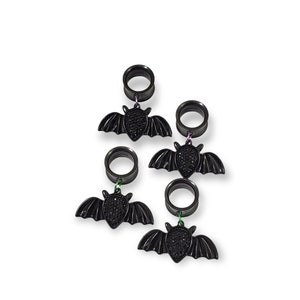 May include: A set of three black ear tunnels, each featuring a dangling bat charm. The bat charms have textured, sparkling black bodies and smooth, outstretched wings. Each tunnel is connected to a bat charm by a small, thin metal jump ring.