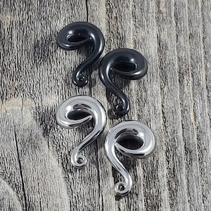 May include: Two pairs of spiral-shaped ear gauges, one pair in a glossy black finish and one pair in a polished silver-tone metal finish. Each gauge features a curved, tapered design that loops into a circular opening, ending in a small point.