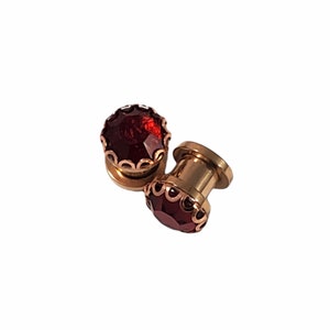 May include: Two gold-colored ear gauges with a large, faceted, deep red gemstone center. The gauges have a decorative, scalloped edge around the gemstone. The jewelry is set against a white background.
