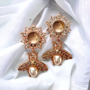 May include: A pair of gold-tone ear gauges with a bee design and a pearl accent. The gauges are adorned with clear rhinestones and have a floral design.