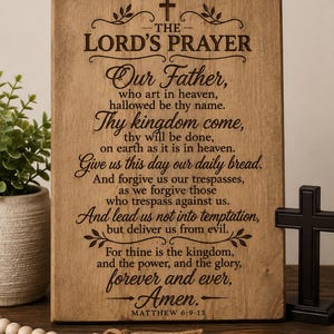 May include: A rectangular light brown wooden sign featuring the text of The Lord's Prayer engraved in dark brown lettering. The sign includes a small cross icon at the top and the citation Matthew 6:9-13 at the bottom, with decorative flourishes.