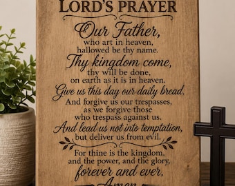 May include: A rectangular light brown wooden sign featuring the text of The Lord's Prayer engraved in dark brown lettering. The sign includes a small cross icon at the top and the citation Matthew 6:9-13 at the bottom, with decorative flourishes.