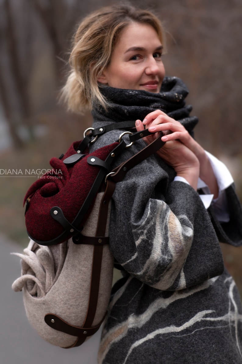 Puede incluir: Una mujer lleva un abrigo gris y blanco con estampado y lleva un bolso de fieltro &uacute;nico con una bolsa de fieltro burdeos adjunta. El bolso tiene una correa de cuero marr&oacute;n y herrajes plateados. El bolso est&aacute; dise&ntilde;ado con una forma org&aacute;nica &uacute;nica.
