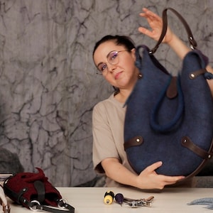 Puede incluir: Una mujer sostiene un bolso tote de fieltro azul oscuro con detalles y correas de cuero marr&oacute;n. El bolso tiene una forma redondeada y est&aacute; hecho de lana felpuda. La mujer lleva gafas y una camisa beige.