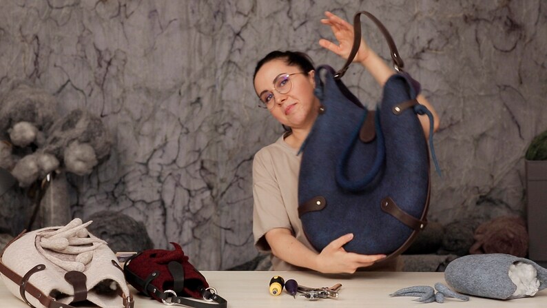 Puede incluir: Una mujer sostiene un bolso tote de fieltro azul oscuro con detalles y correas de cuero marr&oacute;n. El bolso tiene una forma redondeada y est&aacute; hecho de lana felpuda. La mujer lleva gafas y una camisa beige.