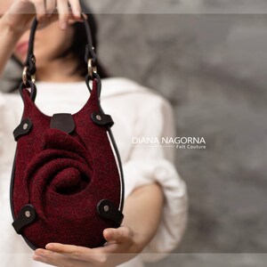 Puede incluir: Un bolso de mano de fieltro burdeos con un dise&ntilde;o de rosa y detalles en cuero negro. El bolso tiene un asa superior y una correa para el hombro.