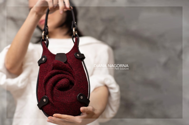 Puede incluir: Un bolso de mano de fieltro burdeos con un dise&ntilde;o de rosa y detalles en cuero negro. El bolso tiene un asa superior y una correa para el hombro.