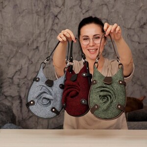 Puede incluir: Tres bolsos de fieltro con adornos en forma de rosa. Los bolsos son de color gris, burdeos y verde.