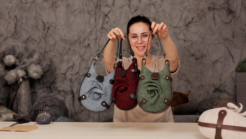 Puede incluir: Tres bolsos de fieltro con adornos en forma de rosa. Los bolsos son de color gris, burdeos y verde.