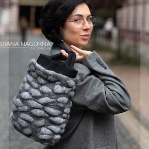 Könnte beinhalten: Eine graue Filmtasche mit einem strukturierten, kieselsteinartigen Design. Die Tasche hat einen schwarzen oberen Teil und einen schwarzen Riemen. Die Tasche wird von einer Person getragen, die einen grauen Mantel und eine Brille trägt. Der Text "DIANA NAGORNA - Felt Couture" ist im Bild sichtbar.