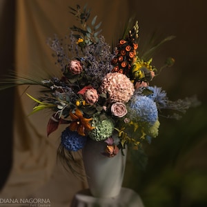 May include: A large, multi-colored floral arrangement in a tall, smooth, light-colored vase. The bouquet features a variety of textures and colors, including pink, blue, green, and orange flowers, along with dried grasses and foliage.