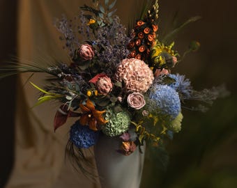 May include: A large, multi-colored floral arrangement in a tall, smooth, light-colored vase. The bouquet features a variety of textures and colors, including pink, blue, green, and orange flowers, along with dried grasses and foliage.