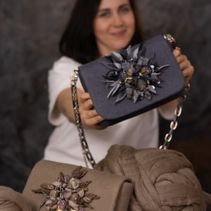 May include: Two decorative handbags with floral embellishments. One is a slate gray color with a silver chain strap, and the other is a tan color. Both bags have a flap closure and are decorated with a cluster of fabric and bead accents.
