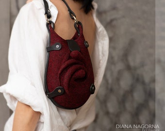 May include: A burgundy felt handbag with a rose-shaped design and black leather accents. The bag has a black leather strap and gold-toned hardware.  DIANA NAGORNA Felt Couture.