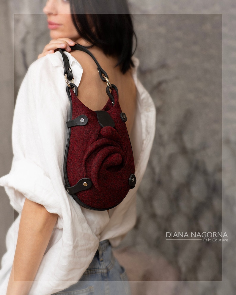 Puede incluir: Un bolso de mano de fieltro burdeos con un dise&ntilde;o en forma de rosa y detalles en cuero negro. El bolso tiene una correa de cuero negro y herrajes dorados. DIANA NAGORNA Felt Couture.