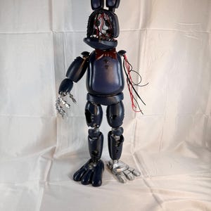 May include: A dark blue rabbit-like animatronic figure with a missing face plate revealing internal wiring. It features a red bowtie, two long ears, a metallic silver hand, and a matching silver foot. The body shows signs of wear with holes.