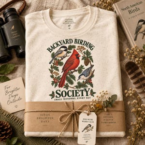 May include: A cream-colored speckled t-shirt featuring a graphic of a red cardinal, a chickadee, and a nuthatch surrounded by pine branches and berries. The text reads BACKYARD BIRDING SOCIETY SMALL WONDERS, EVERY DAY. The shirt is wrapped in a brown paper band.