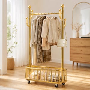 Pode incluir: Um cabideiro de metal dourado com uma barra superior para pendurar com vários ganchos, uma cesta de armazenamento inferior e quatro rodízios. Várias peças de vestuário, incluindo um casaco bege, uma jaqueta marrom estampada e um suéter creme, estão penduradas.