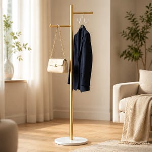 Pode incluir: Um cabideiro de metal dourado com base circular branca, apresentando dois braços horizontais em alturas diferentes. Uma bolsa cor de creme com alça de corrente dourada está pendurada em um braço e um blazer azul-marinho no outro.