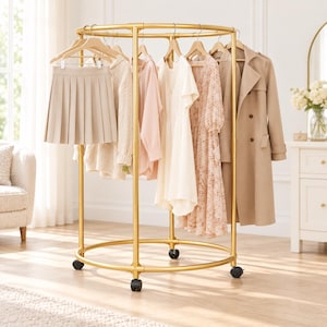 Puede incluir: Un perchero circular de metal dorado con ruedas, que sostiene varias prendas, incluyendo una falda plisada beige, un suéter de punto, una blusa rosa claro, un vestido blanco, un vestido estampado y una gabardina beige en perchas.