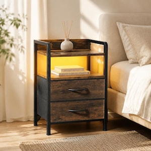 May include: A nightstand with a black metal frame and two drawers featuring a wood grain finish. The unit includes an open shelf with integrated LED lighting and a power outlet. On the shelf sit two books titled Architectural Digest and Vanity Fair 100 Years, topped by a small white vase with wooden reeds.
