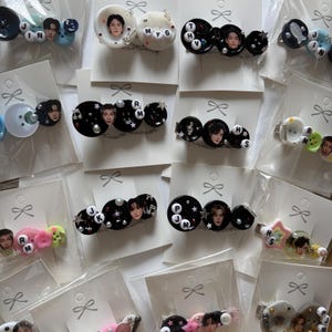 May include: A collection of hair clips featuring circular buttons, small letter beads, and miniature portraits of individuals. The clips are decorated with various charms including stars and musical notes, each mounted on individual white display cards.