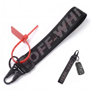 May include: A black fabric keychain strap featuring the white text OFF-WHITE with red stitching details. It includes a black metal lobster clasp, a red plastic zip tie security tag, and a smaller matching keychain attached to a black car key fob.