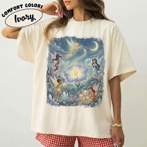 May include: An ivory t-shirt featuring a graphic print of multiple fairies surrounding a large, glowing blue flower. The design includes small mushrooms, crystals, and a crescent moon in the night sky. Text at the top reads COMFORT COLORS Ivory.