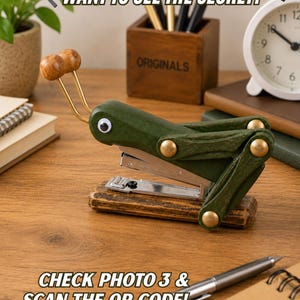 May include: A green grasshopper-shaped stapler with a googly eye, two wooden antennae, and brass-colored rivets. The stapler is mounted on a dark wooden base. Text on the image reads: WANT TO SEE THE SECRET? and CHECK PHOTO 3 & SCAN THE QR CODE!
