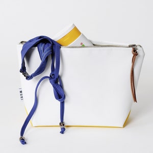 Pu&ograve; includere: Una pochette con zip bianca e gialla con una tracolla in tessuto blu e un tiretto in pelle marrone. La pochette ha la scritta "MURI" stampata sul lato.