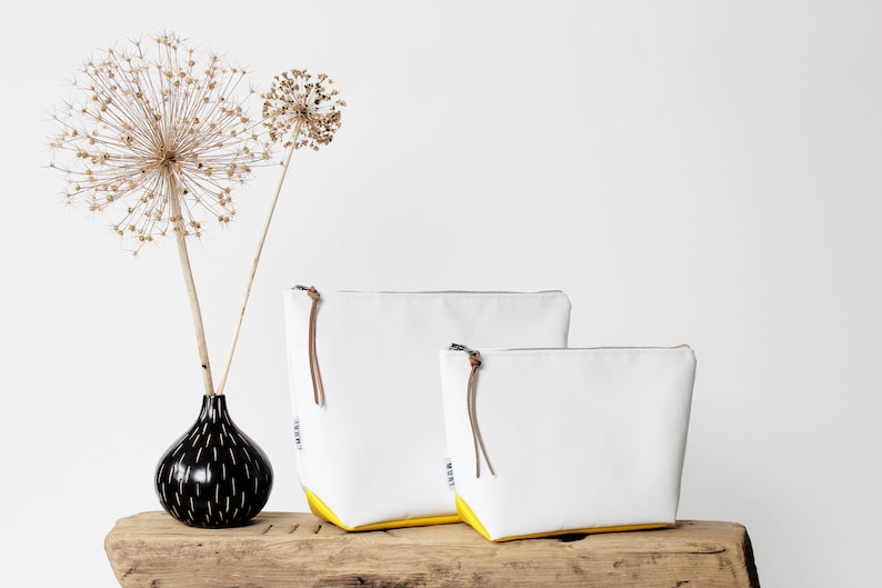 Pu&ograve; includere: Due pochette con cerniera bianche con bordino giallo, una pi&ugrave; grande dell'altra, appoggiate su una superficie di legno. Un vaso nero con un motivo bianco contiene fiori secchi.