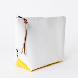 Pu&ograve; includere: Borsa a zip in tela bianca con fondo giallo e tiretto in pelle marrone. La borsa ha un'etichetta bianca con la parola "MOUTHI" stampata su di essa.