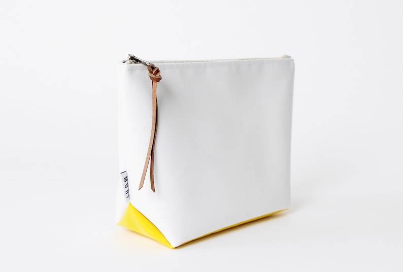 Pu&ograve; includere: Borsa a zip in tela bianca con fondo giallo e tiretto in pelle marrone. La borsa ha un'etichetta bianca con la parola "MOUTHI" stampata su di essa.