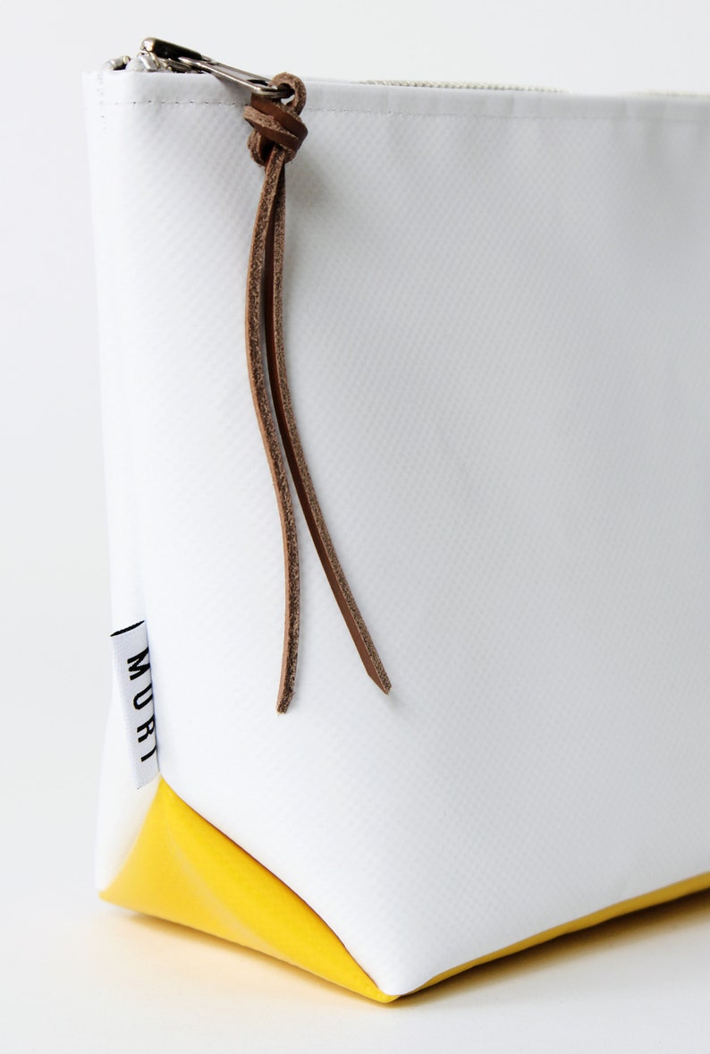 Pu&ograve; includere: Borsa bianca con cerniera, fondo giallo e cinturino in pelle marrone. La borsa ha la scritta "MURI" stampata sul lato.
