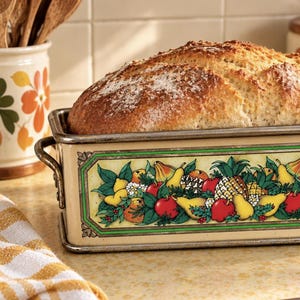 May include: A rectangular metal loaf pan with a cream-colored finish and a decorative border featuring a colorful illustration of various fruits including apples, pears, and pineapples, containing a freshly baked loaf of bread.