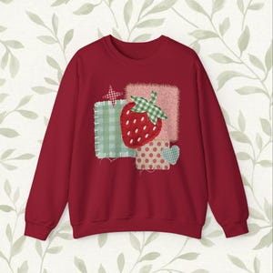 May include: A deep red crewneck sweatshirt featuring a central graphic design of a red strawberry with a green checkered leaf. The strawberry is layered over various fabric-like patches including green gingham, red polka dots, and a small heart.