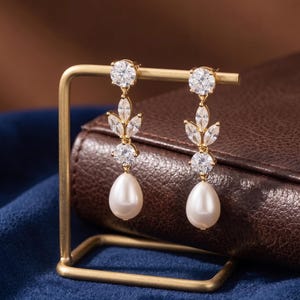 May include: A pair of gold-tone drop earrings featuring a round clear stone at the top, followed by a cluster of three marquise-cut clear stones arranged in a leaf pattern, another round clear stone, and a smooth white teardrop-shaped pearl.