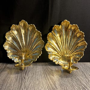 May include: A pair of gold-colored metal wall sconces, each featuring a large, fan-shaped shell backplate with deep vertical ridges and a circular candle holder platform extending from the bottom center of the shell design.
