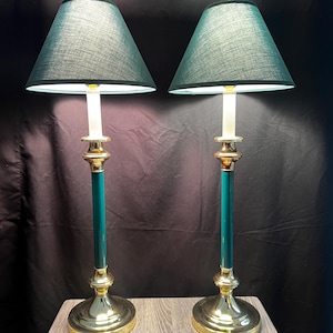 May include: A pair of tall table lamps featuring polished gold-tone metal bases and stems with dark teal accents. Each lamp is topped with a conical, dark teal fabric shade that emits a soft glow from the light source positioned underneath.
