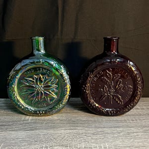 May include: Two round glass bottles with iridescent finishes. The green bottle features a poinsettia design with the text CHRISTMAS 1974. The dark red bottle features a holly design with the text CHRISTMAS HOLLY 1973. Both have narrow necks.