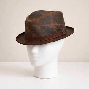 Può includere: Un cappello fedora marrone scuro con una texture vissuta e usurata, caratterizzato da una fascia marrone liscia e solida che circonda la base della corona. Il cappello presenta una corona pizzicata e una tesa strutturata dai toni terrosi.