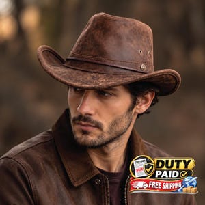 Könnte beinhalten: Ein brauner Leder-Cowboyhut mit Used-Optik, ausgestattet mit einem schmalen, passenden Lederband um die Krone und kleinen Belüftungsösen aus Metall an den Seiten. Der Hut hat eine strukturierte Krempe und ein rundes Metallemblem am Hutband.