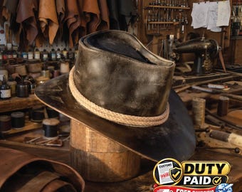 May include: A dark brown leather hat with a wide brim and a light tan braided rope band around the crown. The hat is positioned on a wooden stand, showcasing a distressed finish and a structured shape with a deep indentation on the top crown.
