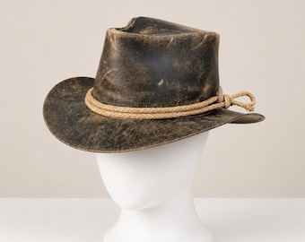 May include: A dark brown, distressed leather hat with a weathered finish and a pinched crown. The hat features a wide, slightly curved brim and is accented with a thick, light tan braided rope band tied in a simple knot on the right side.